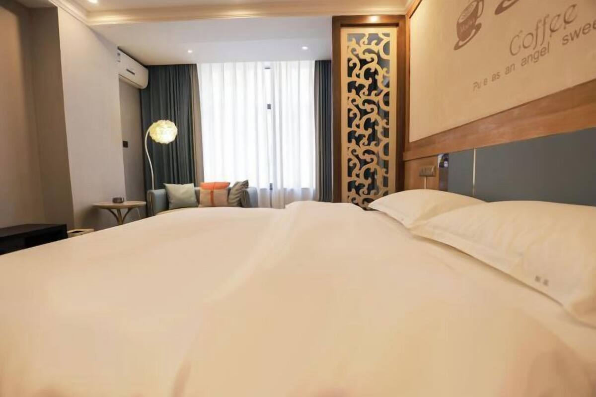 Hotellihuone, jossa on suuri sänky, yöpöytä, lamppu, koristeellinen seinäpaneeli ja ikkuna, jossa on verhot.