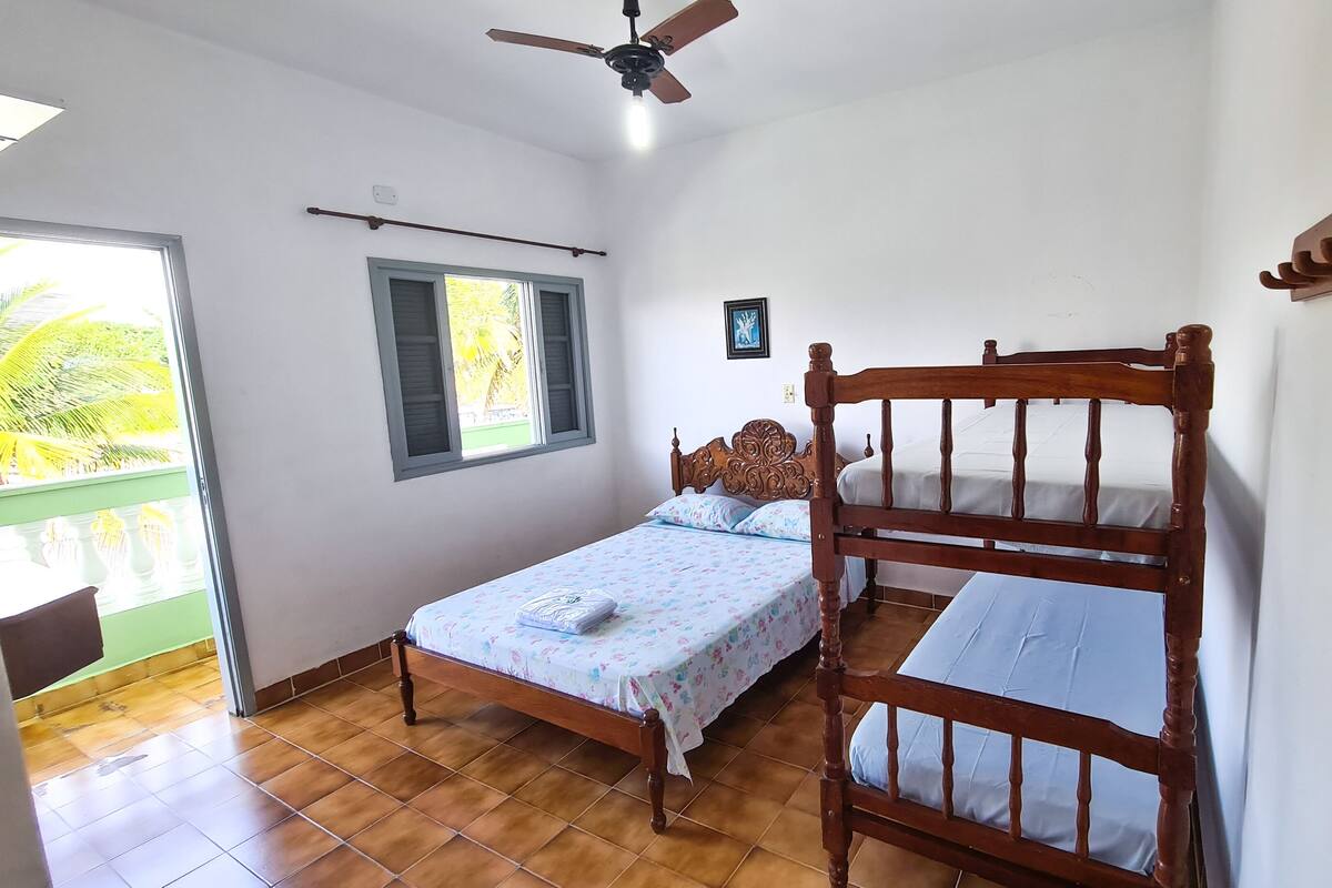 Un dormitorio con una cama individual, cabecera de madera, ventilador de techo, ventana con contraventanas y vista a palmeras al exterior.