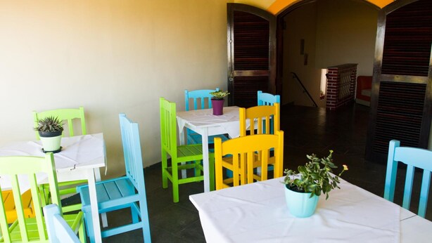 A dining area with colorful chairs and tables, a potted plant on each table, and a brick fireplace in the background. - Hotel Casarao (Peruíbe)