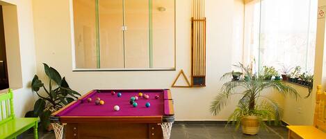 A billiards table with balls, a green bench, and potted plants in a room with large windows and a yellow chair.