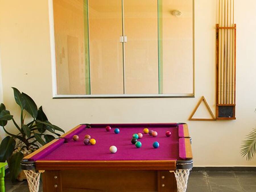 Ein Billardtisch mit Kugeln, eine grüne Bank und Topfpflanzen in einem Raum mit großen Fenstern und einem gelben Sessel.