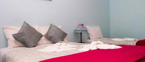 Ein ordentlich bezogenes Bett mit floralem Überwurf und einer roten Decke, Kissen und ein kleiner Tisch mit einer Lampe.