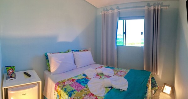 A bedroom with a bed, pillows, a nightstand, a remote control, and a window with curtains. - Hotel Casarao (Peruíbe)