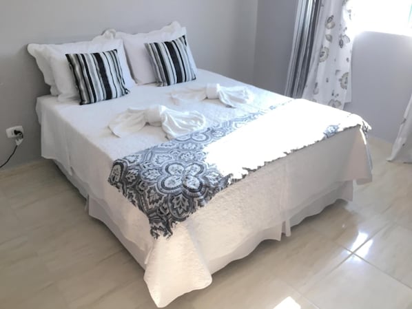 Una cama bien hecha con sábanas blancas y almohadas a rayas, una manta estampada y un enchufe en la pared.