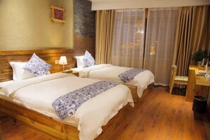 Habitación de hotel con dos camas, cabeceros de madera, un escritorio pequeño con una silla, un cubo de basura y una ventana con cortinas.