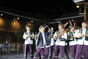 Un grupo de personas vestidas con trajes tradicionales actuando en un escenario, con un edificio de madera al fondo.