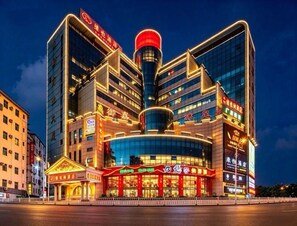 A multi-story hotel with a prominent central tower, multiple signage, and a well-lit facade. - Vienna Hotel (Dongguan)