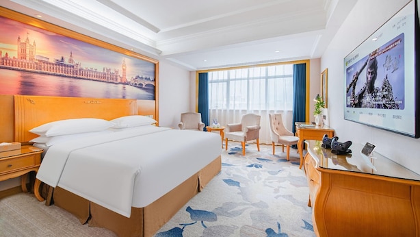A hotel room with a large bed, a desk, a chair, a TV, and a painting of a cityscape. - Vienna Hotel (Dongguan)