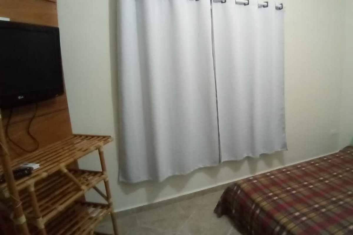A small room with a bed, a television on a wooden shelf, and white curtains.