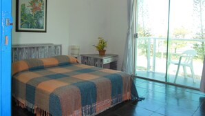 A bedroom with a bed, a desk, a chair, a potted plant, and a balcony with a view of trees. - Praia Sul Studios (Peruíbe)