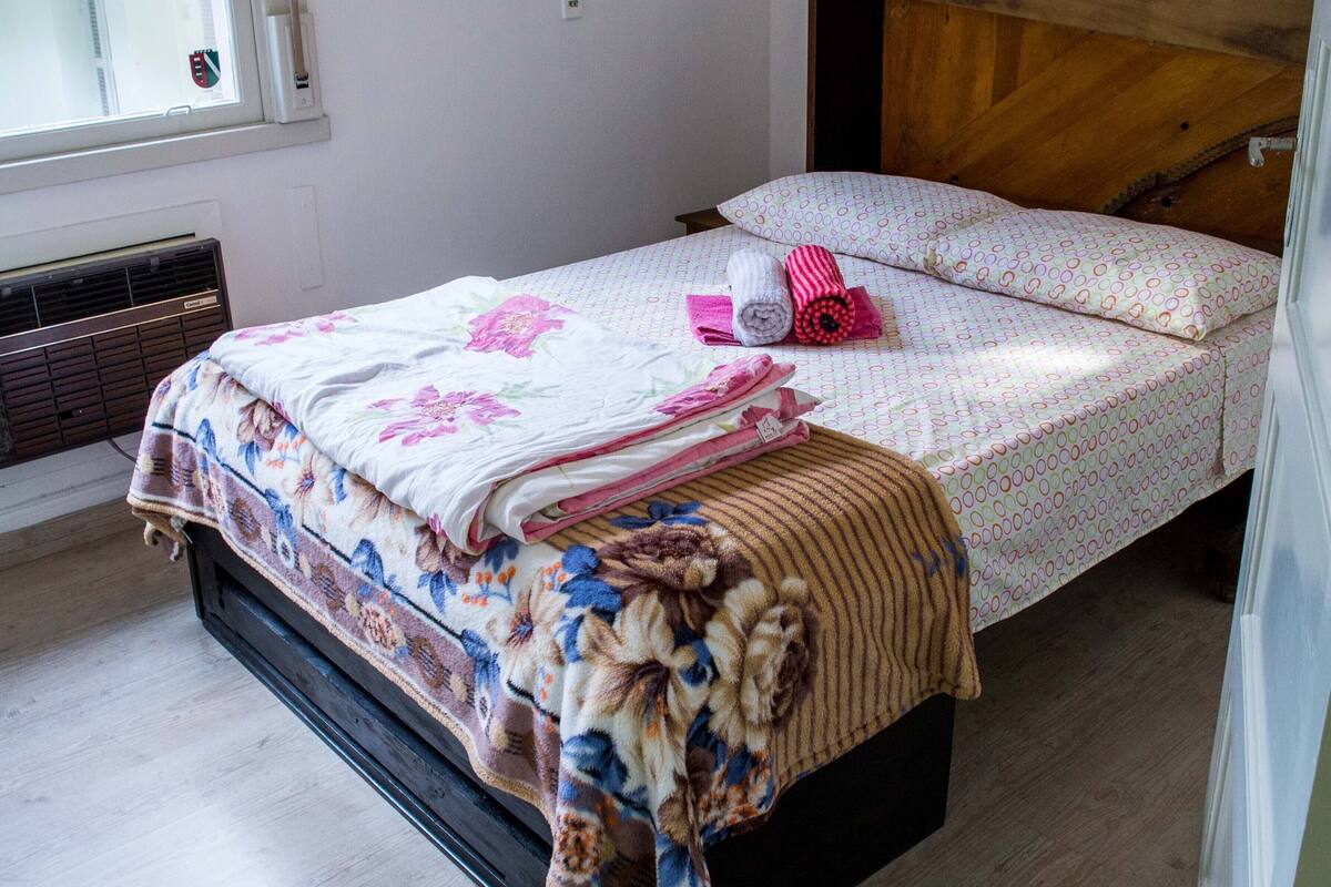 Een net opgemaakt bed met bloemenbeddengoed, een houten hoofdbord en een raam met jaloezieën.
