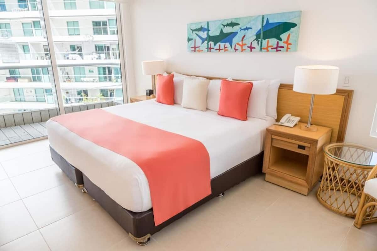 Quarto de hotel com uma cama grande, cabeceira de madeira, mesa de cabeceira com abajur, cadeira de vime e vista de um edifício através de uma janela ampla.