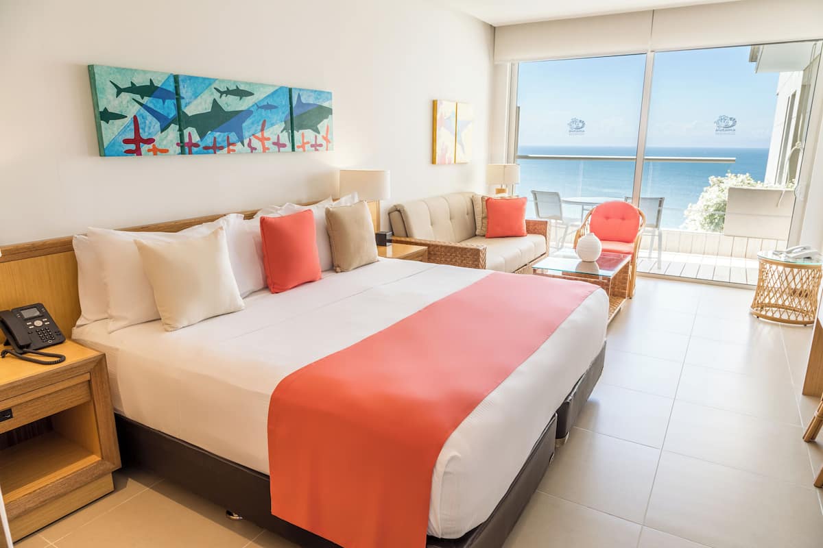 Quarto de hotel com uma cama grande, um sofá, uma mesinha e vista para o mar.