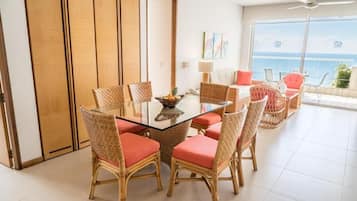 Un comedor con mesa de vidrio y sillas de mimbre, un sofá con cojines, y vista al mar a través de amplios ventanales.