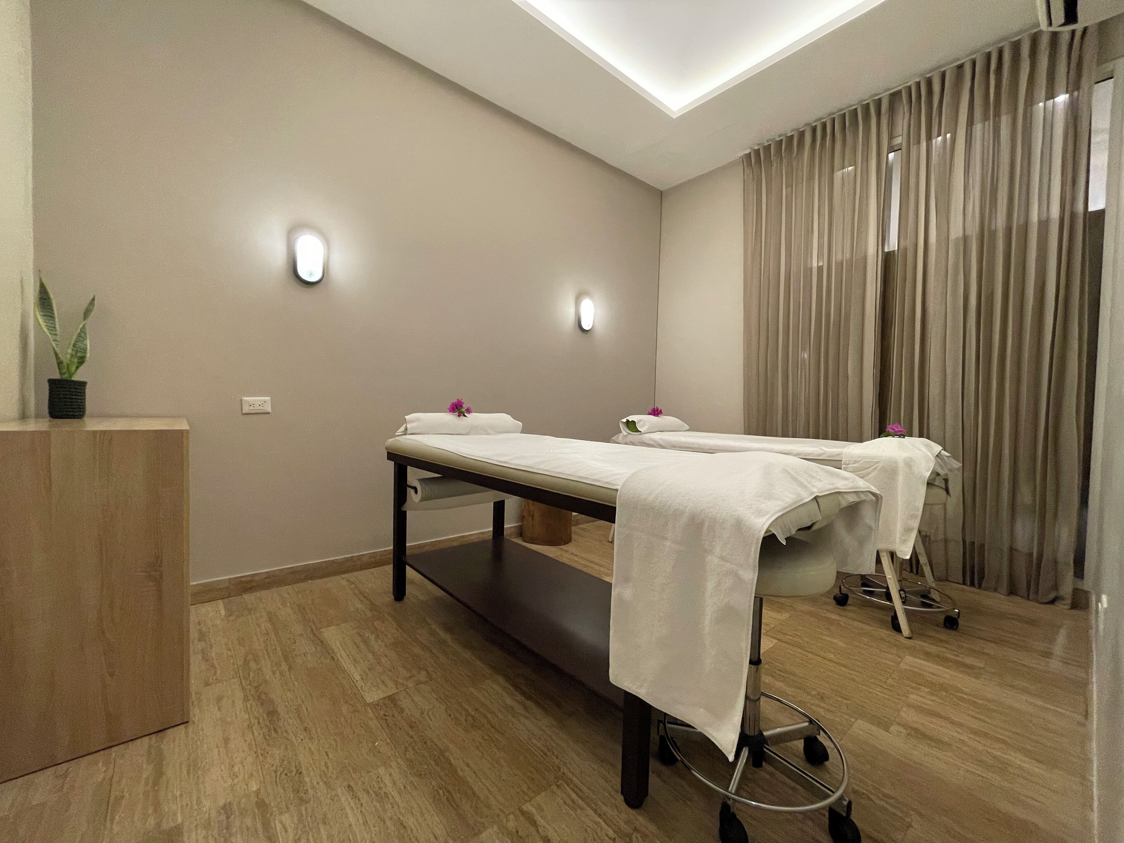 A modern spa room with two massage tables, a wooden cabinet, and a potted plant.