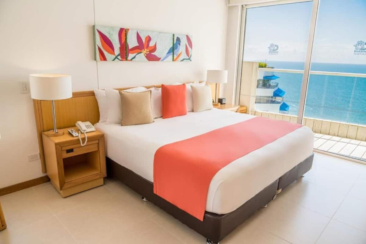 Quarto de hotel com cama, luminárias de cabeceira, telefone, pintura e varanda com vista para o mar.