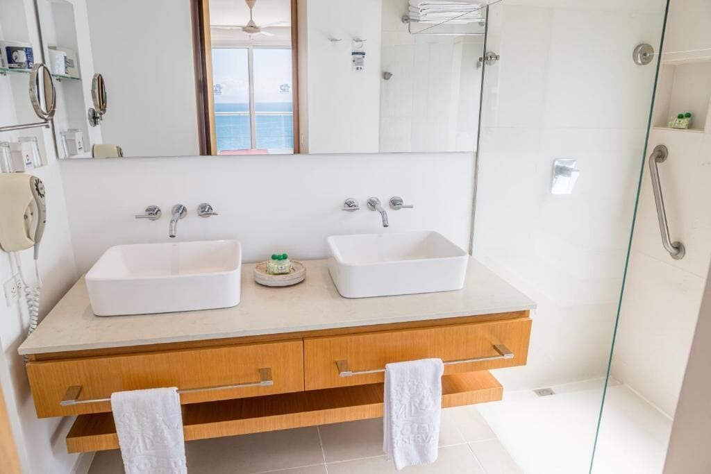 A modern bathroom with two sinks, a vanity with drawers, a glass shower enclosure, and a view of the sea through a window.