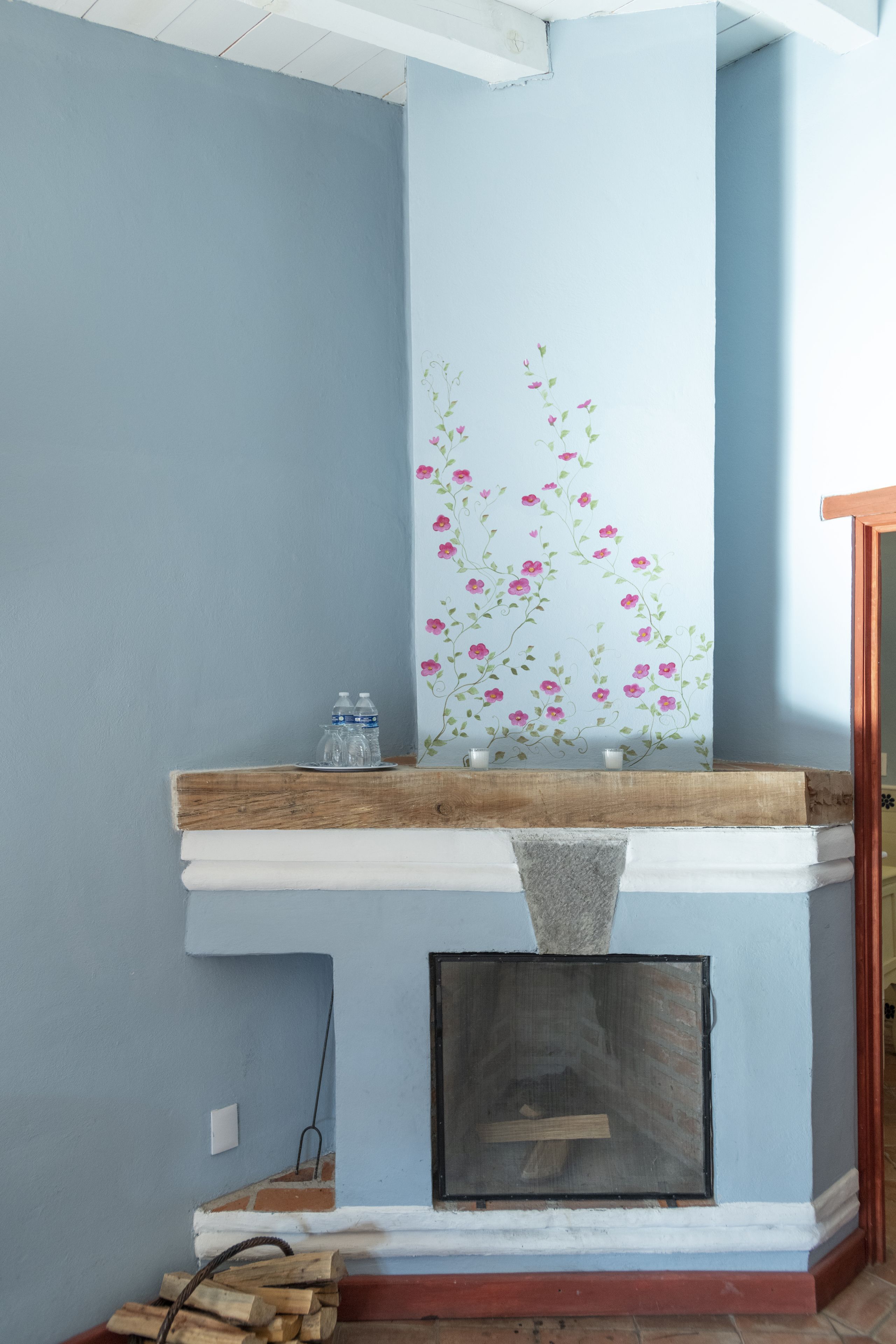 Una chimenea con repisa blanca, un estante pequeño con botellas de agua y un mural floral decorativo.