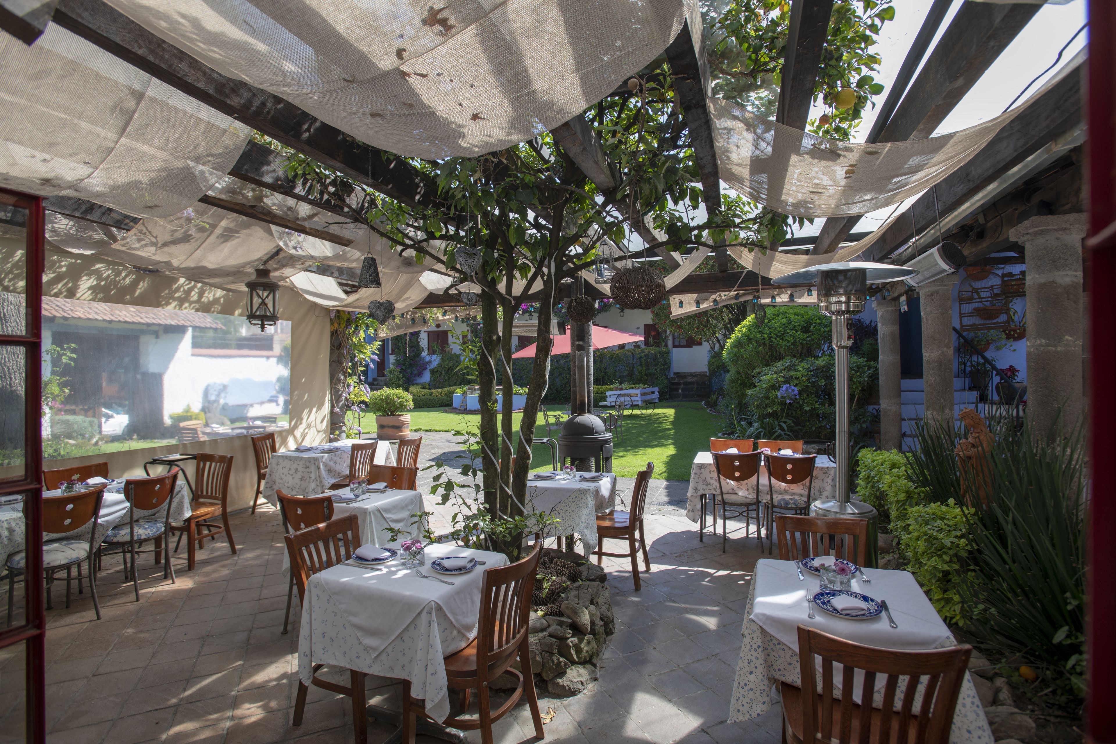 An outdoor patio area with tables and chairs, covered by a canopy, and surrounded by greenery.