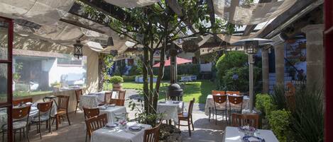 An outdoor patio area with tables and chairs, covered by a canopy, and surrounded by greenery.