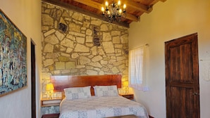 A bedroom with a stone wall, wooden ceiling, a bed with floral bedding, a wooden bench, and a door.