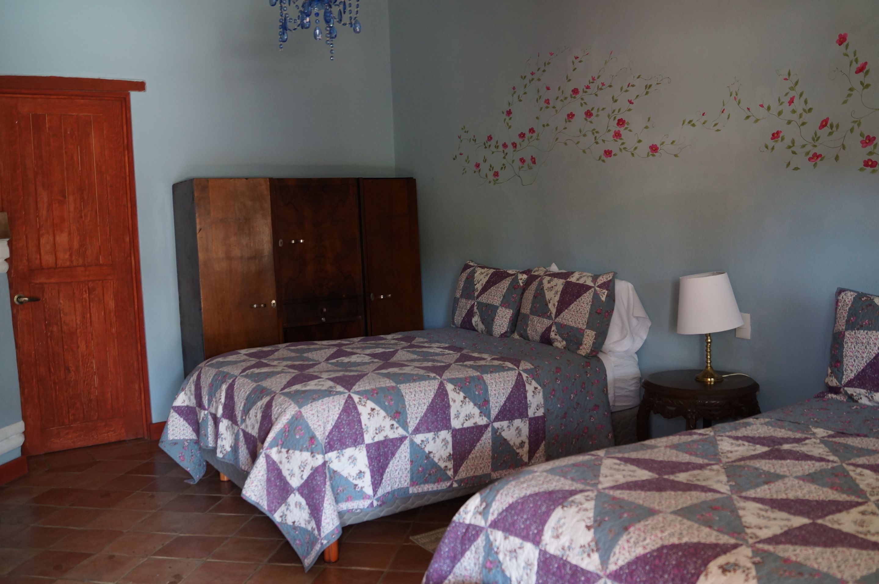 Un dormitorio con dos camas, un armario de madera, una mesita de noche con lámpara y decoración floral en la pared.