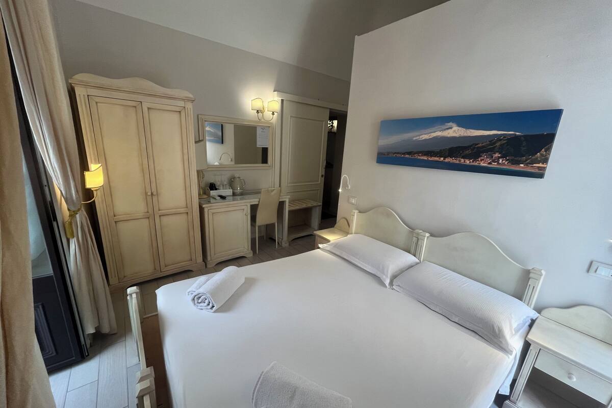 A hotel room with a large bed, a desk with a chair, a wardrobe, and a painting on the wall.