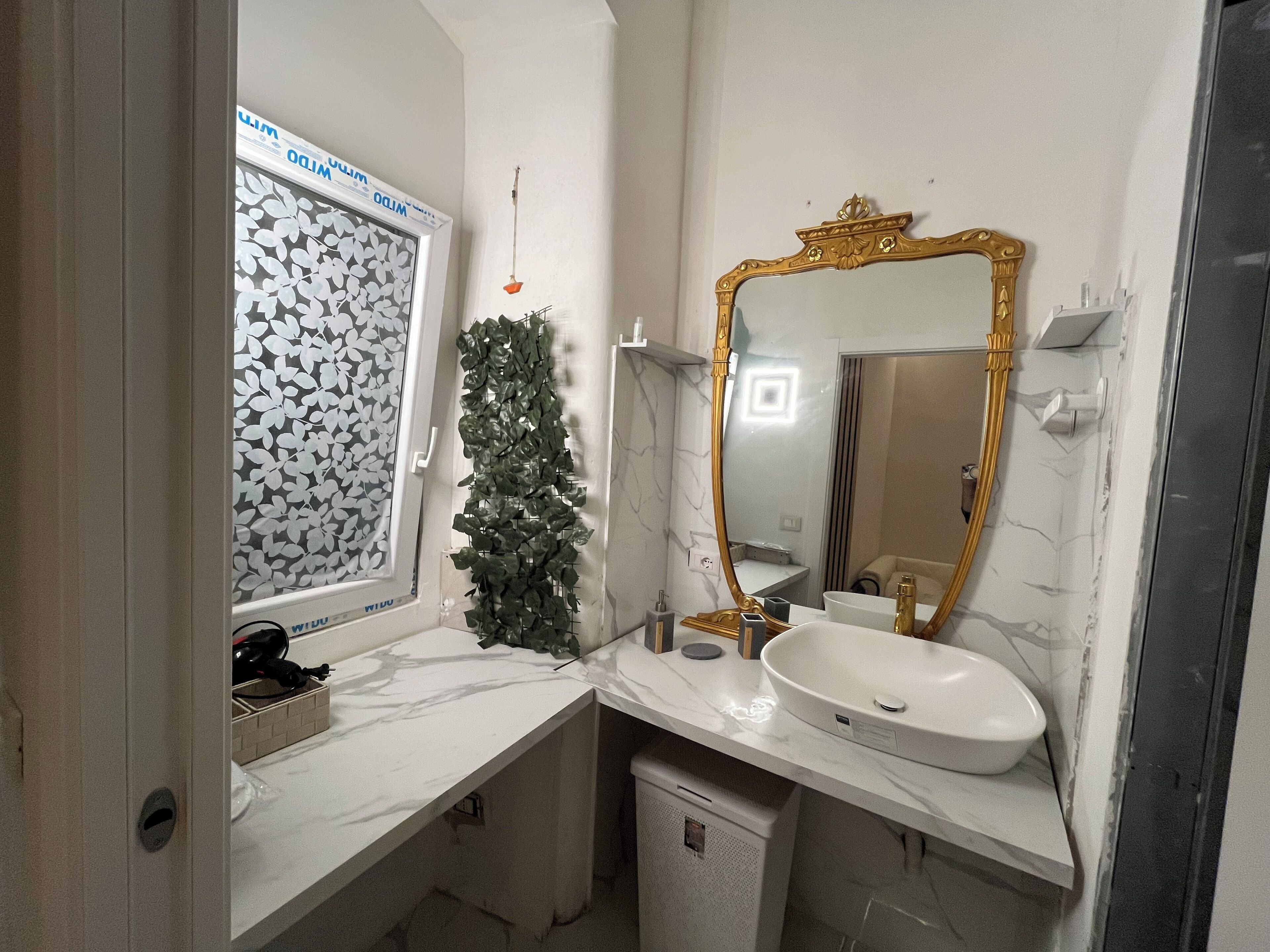 Une salle de bain avec un lavabo blanc, un miroir à cadre doré, une fenêtre avec un store à motifs de feuilles et une petite plante verte.