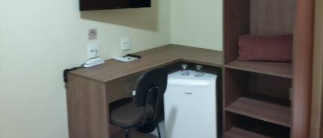 Habitación compacta con escritorio, silla y un electrodoméstico blanco pequeño, probablemente un refrigerador o calefactor. Un televisor está montado en la pared sobre el escritorio.
