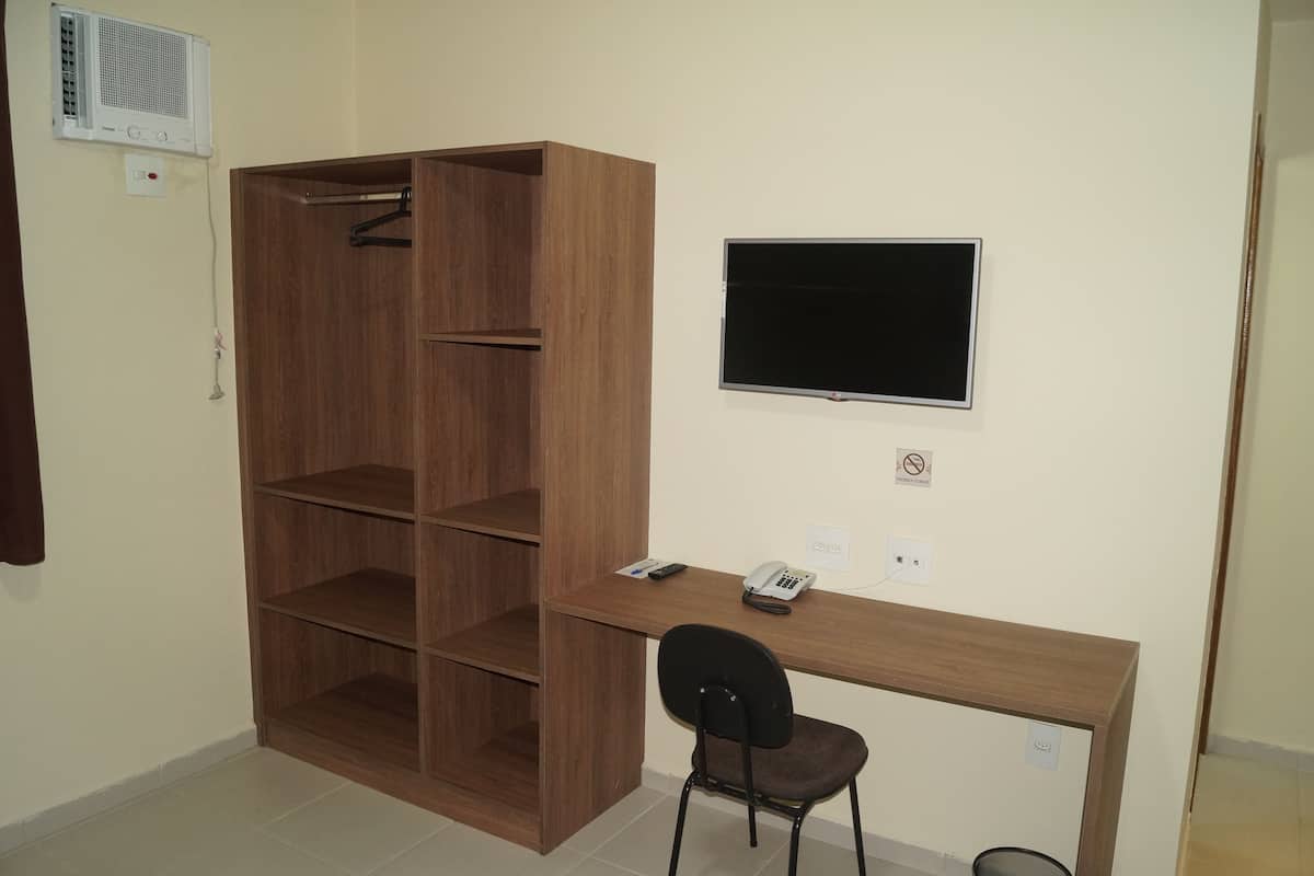 A room with a wooden wardrobe, a desk with a chair, a mounted TV, and a telephone.