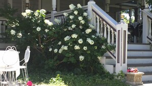 Un edificio con una escalera blanca, barandillas blancas y una puerta blanca. Hay una silla blanca y una planta en maceta con flores rosas en frente del edificio.