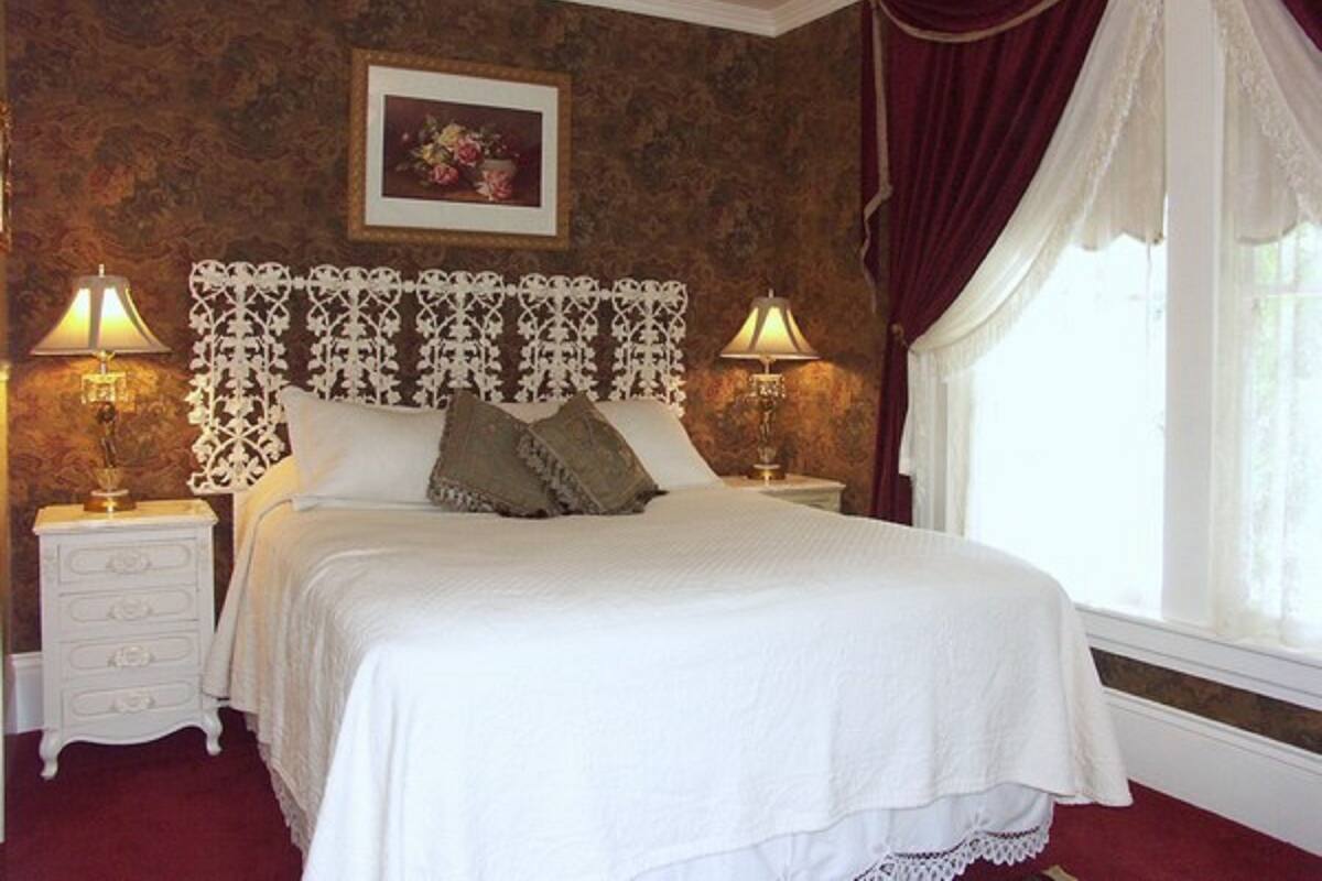 Een slaapkamer met een groot bed, twee nachtkastjes met lampen, een decoratief hoofdeinde, een bloemistisch kunstwerk in een lijst aan de muur, en een raam met gordijnen.