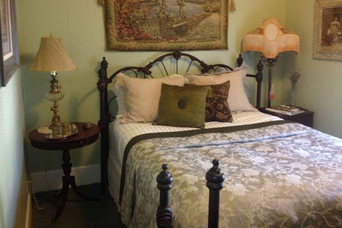 Een slaapkamer met een hemelbed, een nachtkastje met een lamp, een lijst met een schilderij boven het bed en een klein tafeltje met een lamp ernaast.