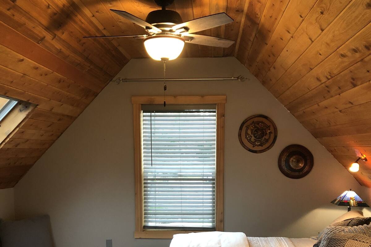 Een knus zolderkamer met een schuine kap, een plafondventilator, een groot raam met zonwering, een bed met een witte dekbedovertrek, een kamerbreed tapijt met dessin en een klein tafeltje met een lamp.