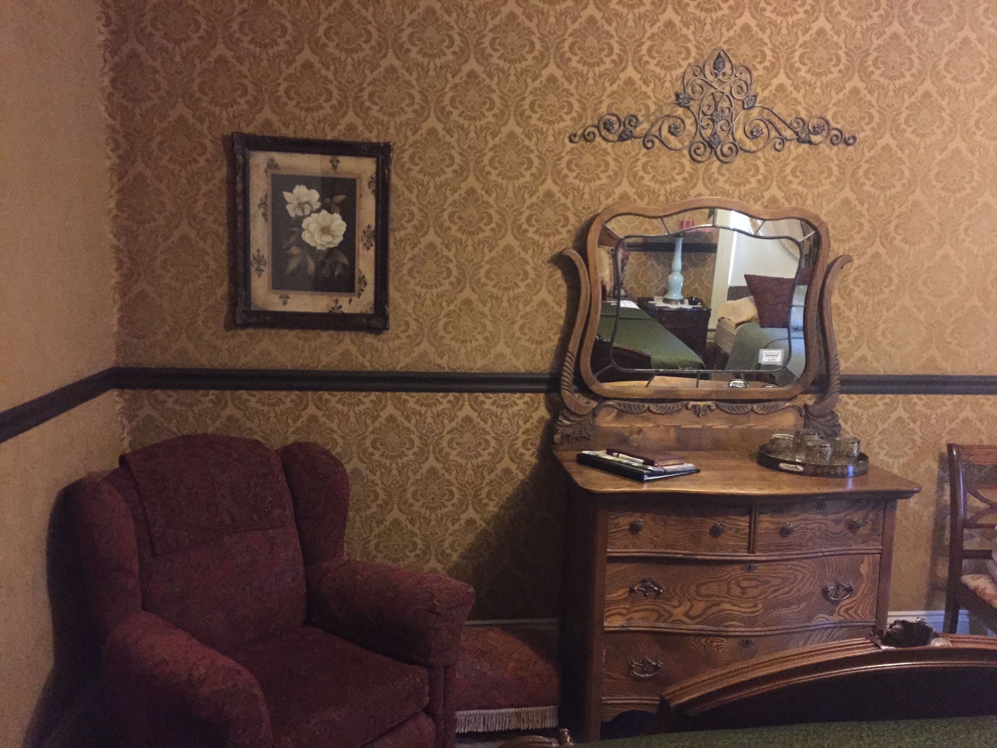 A room with a patterned wallpaper, a wooden dresser with a mirror, a framed picture, and a red armchair.