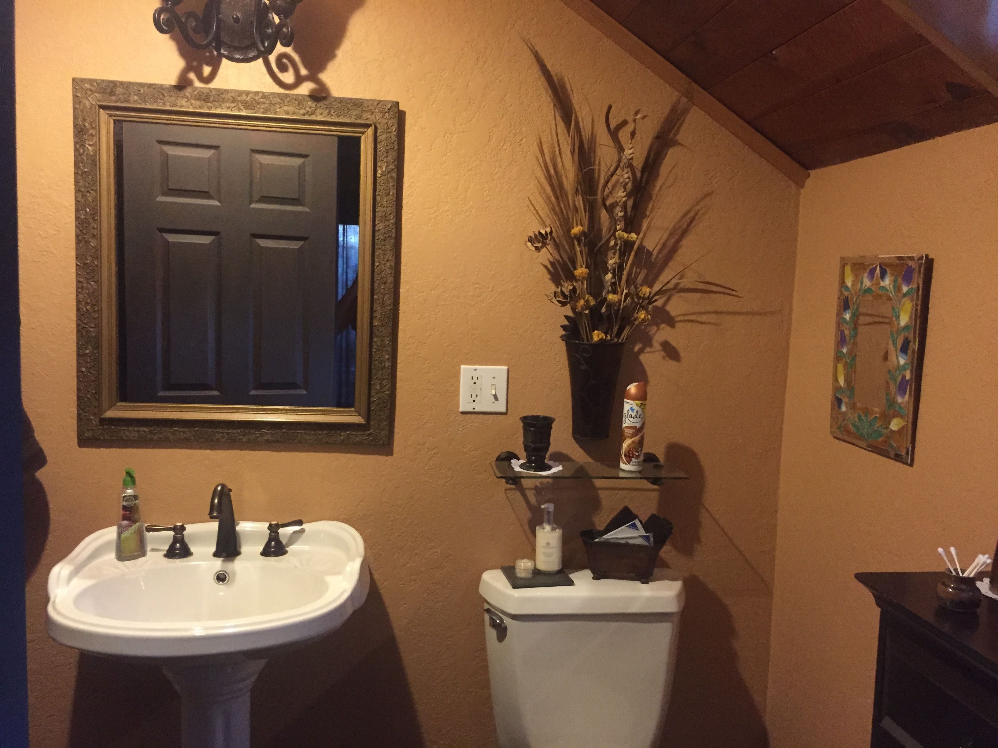 A bathroom with a pedestal sink, a toilet, a framed mirror, a vase with dried plants, and a small shelf with toiletries.