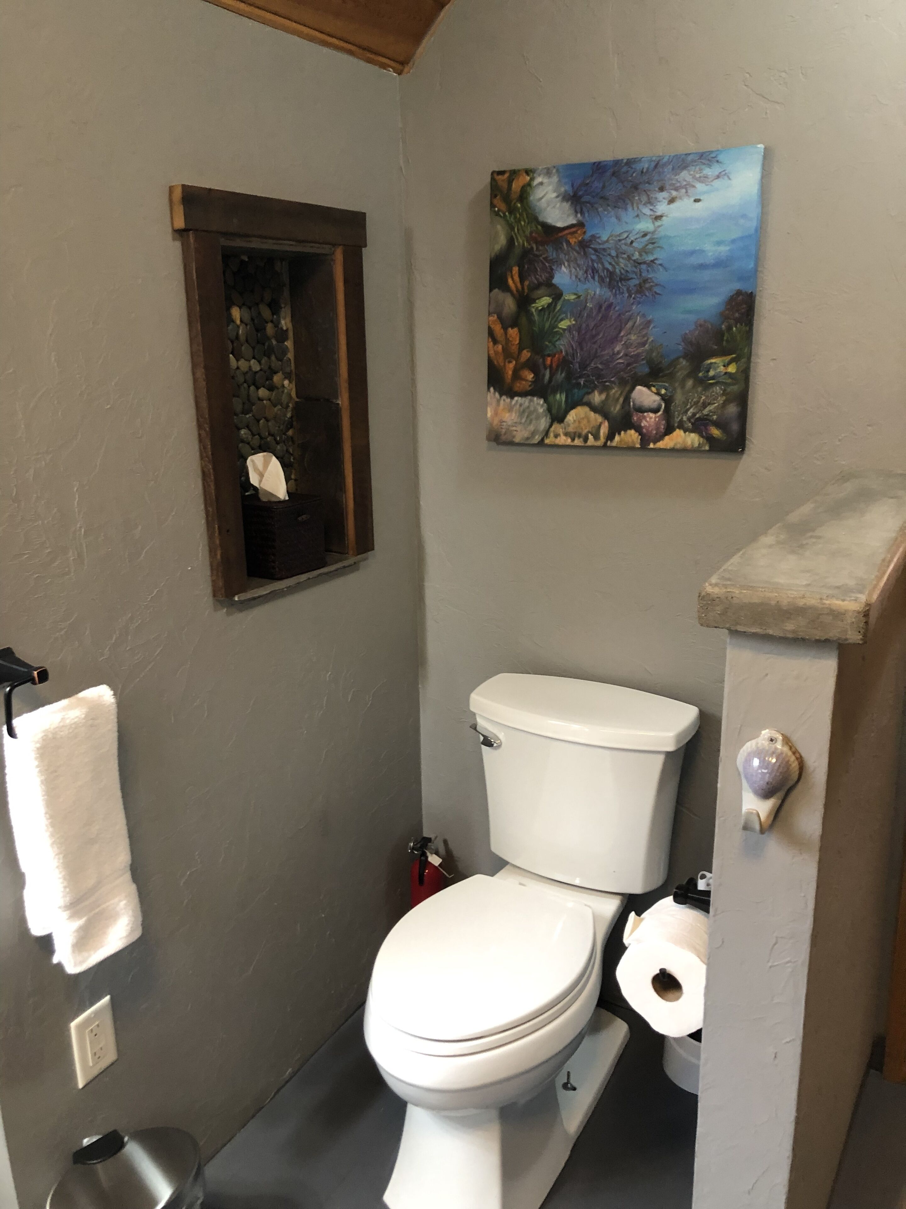 A bathroom with a toilet, a towel rack, a painting of an underwater scene, and a small window with a stone shelf.