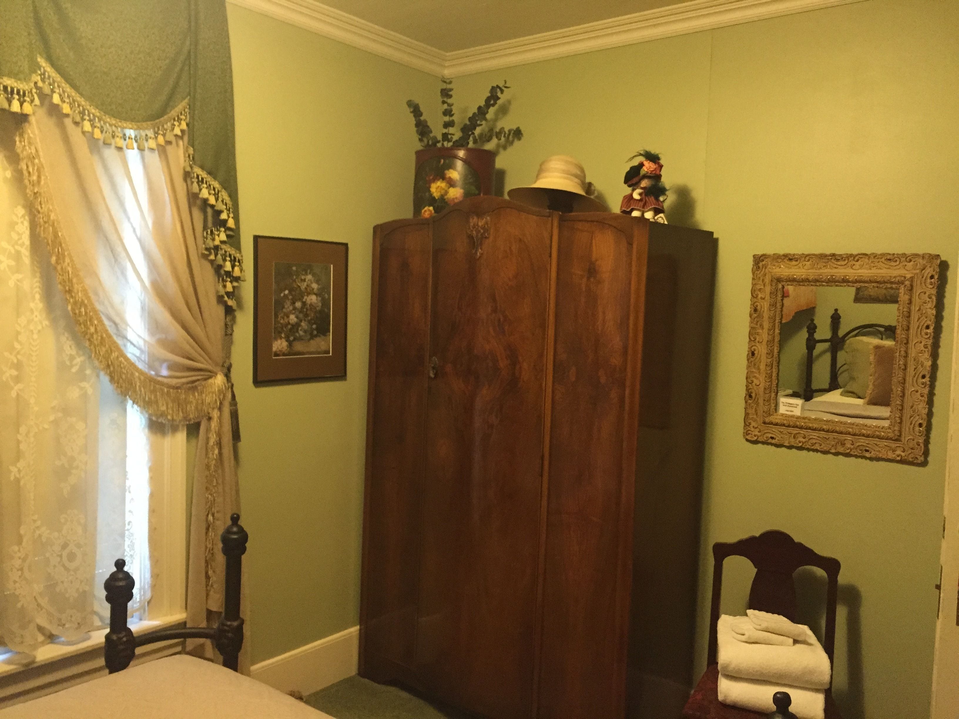 A bedroom with a wooden wardrobe, a bed with a white comforter, a window with sheer curtains, a framed mirror, and a small bedside table with a towel.