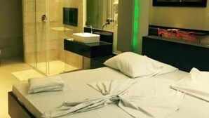 Una habitación de hotel moderna con cabina de ducha de vidrio, cama blanca con toallas ordenadas y una mesita de noche con espejo y una pequeña silla roja.