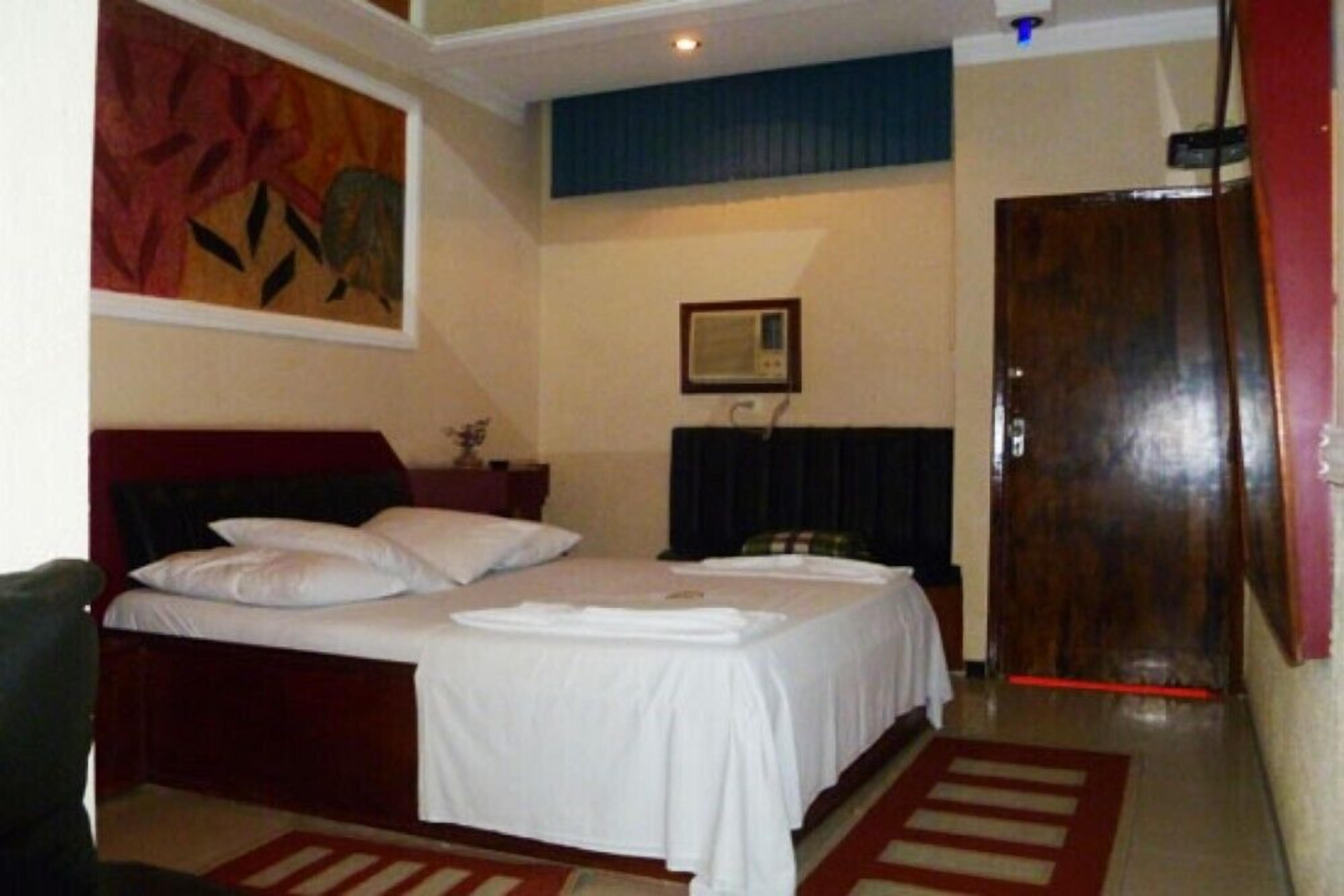 Quarto de hotel com cama de casal, uma mesinha, um quadro na parede e uma porta de madeira.