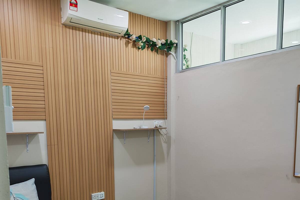A bedroom with a bed, a bedside table, an air conditioning unit, a ceiling fan, a window, and a wooden wall panel.