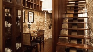 A wooden staircase with a stone wall backdrop, a dining table with chairs, and shelves with various items. - DuCarmo Apartments Caves Vinho do Porto (Vila Nova de Gaia)