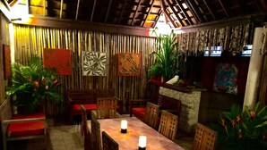 Un espace de restauration intérieur avec une longue table, des chaises et des bougies. Des plantes et des objets décoratifs ornent les murs.
