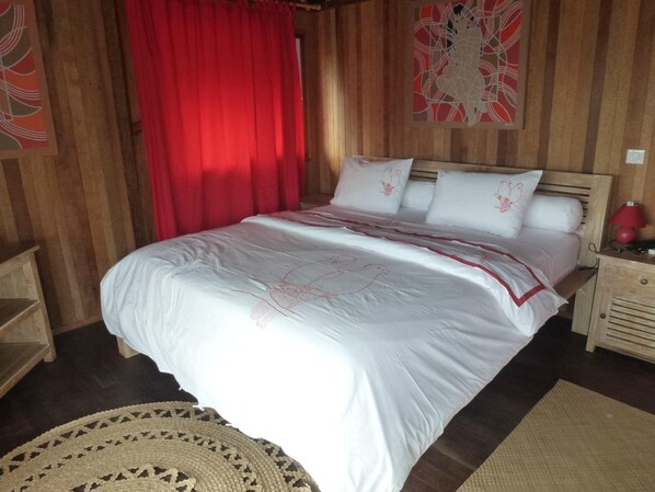 A wooden-paneled room with a bed, red curtains, and a wall painting. - Pension La Perruche Rouge (Rimatara Island)