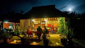 Un espace extérieur aménagé pour les repas, avec des tables et des chaises, entouré de plantes en pot et bordé d’un bâtiment doté d’un toit en pente.