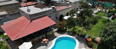 A swimming pool with lounge chairs and umbrellas, surrounded by greenery and a building with a red roof.