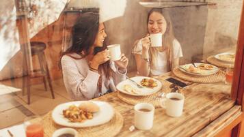 Une table dressée pour le petit-déjeuner, avec deux personnes savourant du café et des viennoiseries, dans un décor rustique et avec vue sur l’extérieur à travers une fenêtre en verre.