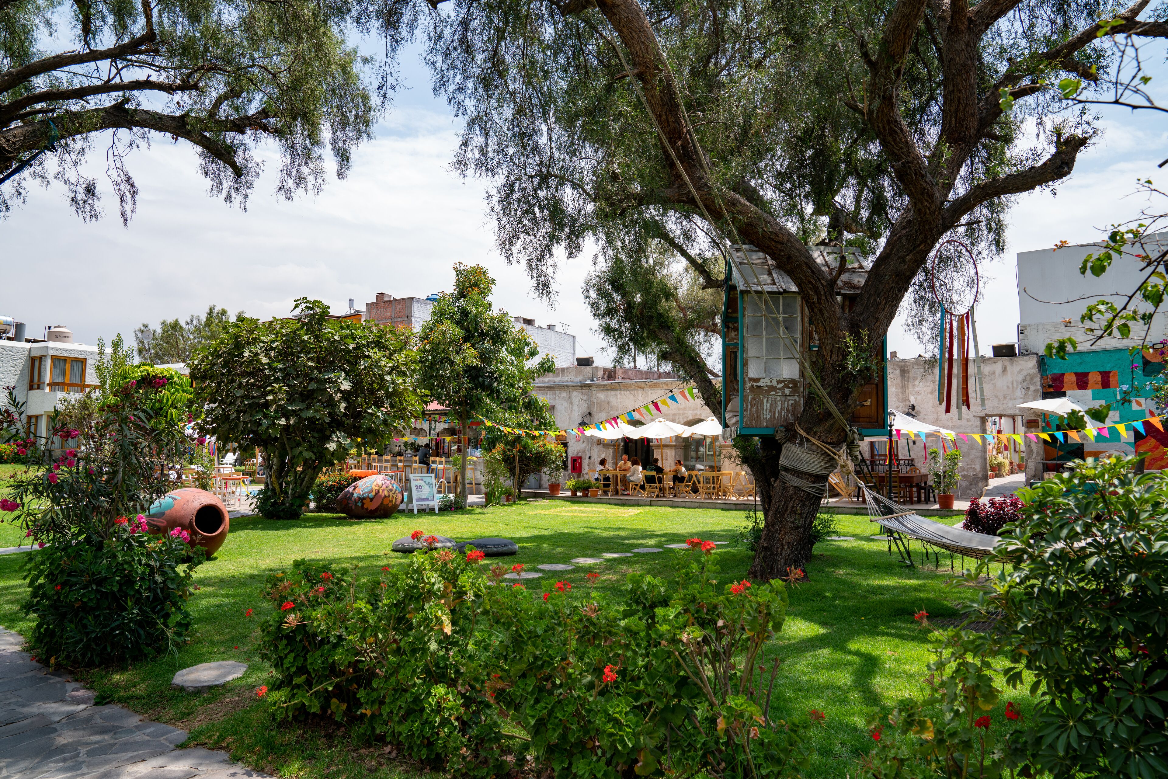 A garden with a large tree, a hammock, and a café area with tables and chairs.