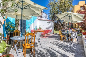 Un patio exterior con mesas y sillas, cubierto por sombrillas y rodeado de plantas en macetas y árboles.