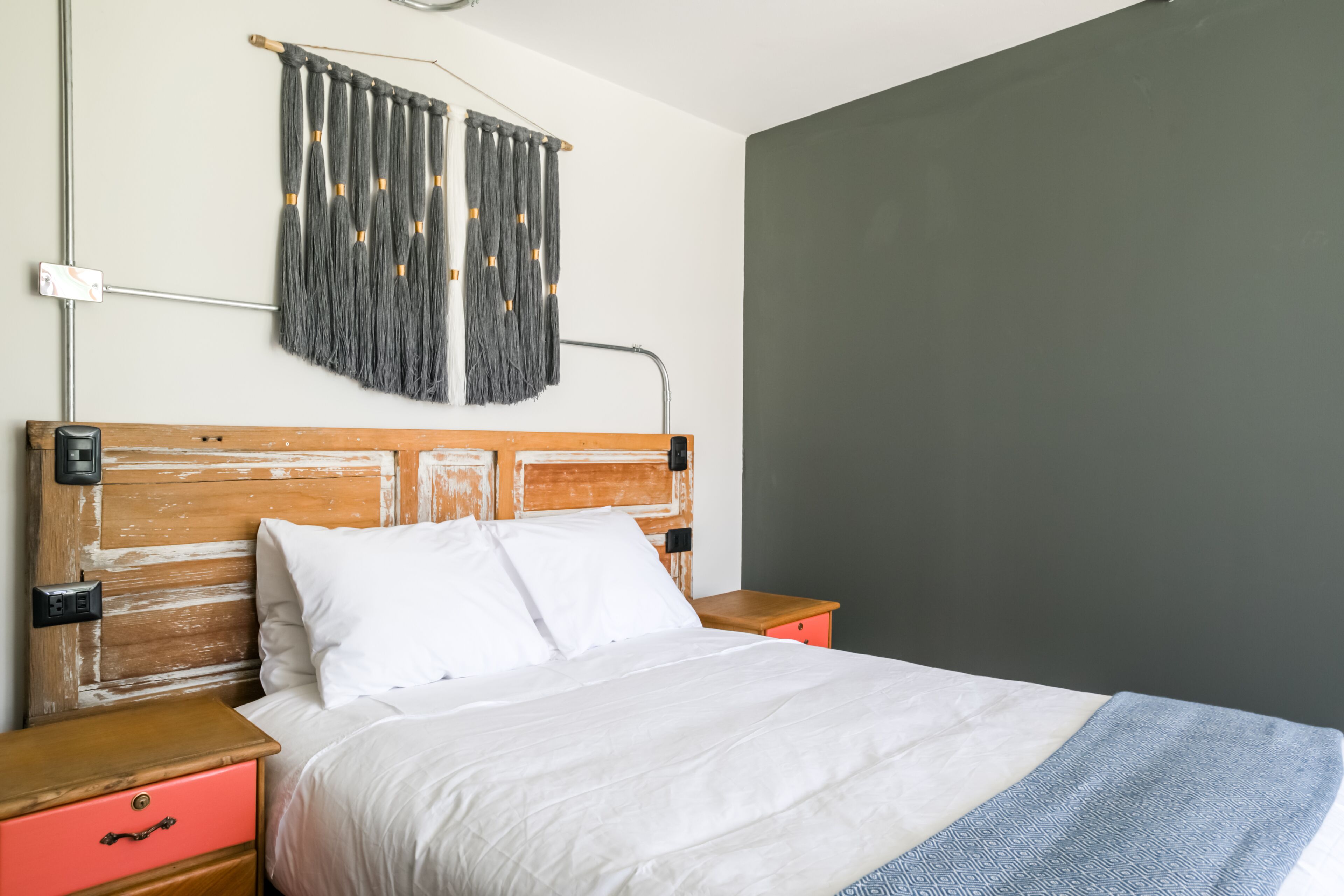 A bedroom with a wooden headboard, white bedding, a red nightstand, and a grey wall.